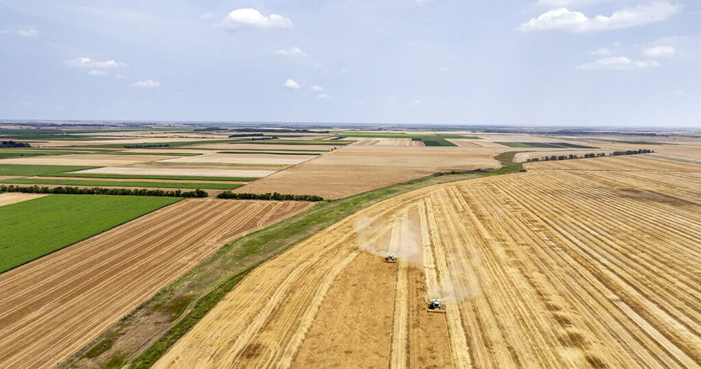 Aerial view of two combine harvesters actively harvesting a vast golden crop field on a large-scale organic farm, with diverse patchwork fields in the background.