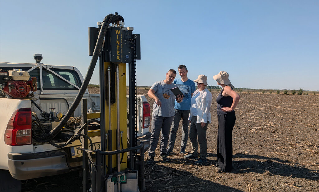 LoginEKO agronomists and Wageningen researchers reviewing sampling data on a tablet beside a Wintex automated soil sampler mounted on a pickup, in a harvested maize field