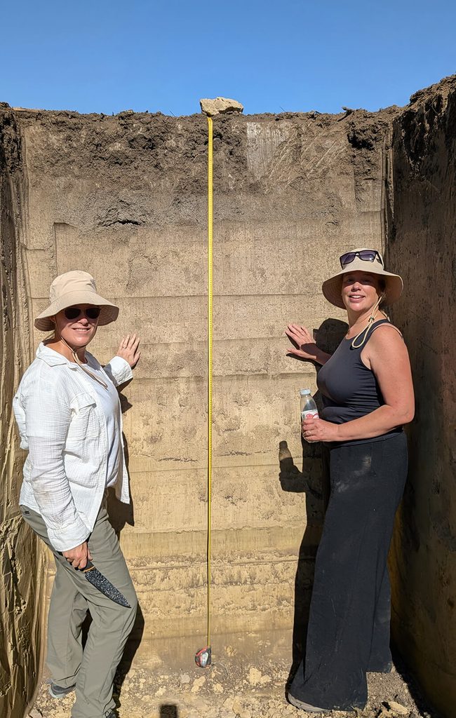 ALT Attribute:
Two soil scientists standing in an open soil profile pit examining horizons with a measuring tape