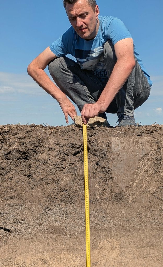 ALT Attribute:
Agronomist crouching at the edge of an open soil pit, holding a tape measure against the exposed profile wall where dark topsoil meets lighter subsoil